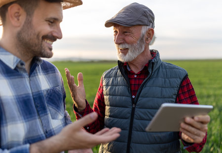 Two smiling farmers standing in green wheat field, using tablet and talking in spring timeの写真素材