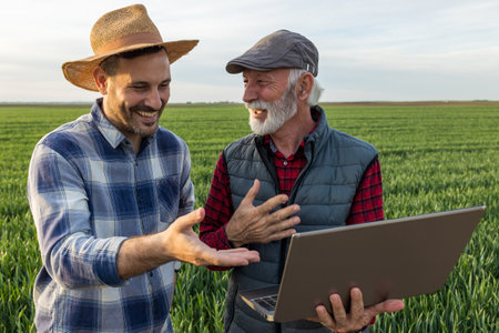 Two farmers standing in green wheat field, using laptop and laughing in spring time