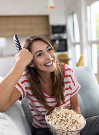 Smiling young woman holding bowl with popcorn and remote and watching movie at home in cozy living roomの写真素材