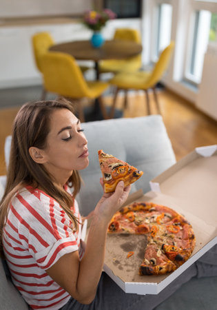 Pretty young woman eating pizza slice on couch in cozy living room at homeの写真素材