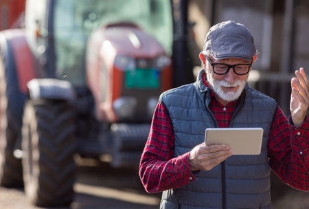 Portrait of excited senior man looking at tablet in front of tractor in cowshed on cattle ranchの写真素材