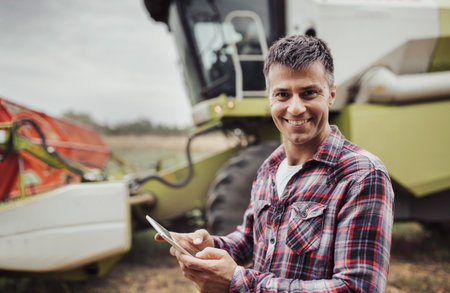 Portrait of handsome farmer holding tablet in front of combine harvester working in field in background, in autumnの写真素材