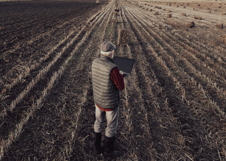 Aerial image of senior farmer standing in field after harvest and baling and working on laptop