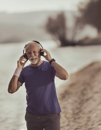 Senior man warming up for jogging on beach and setting headset on ears for motivating music for...
