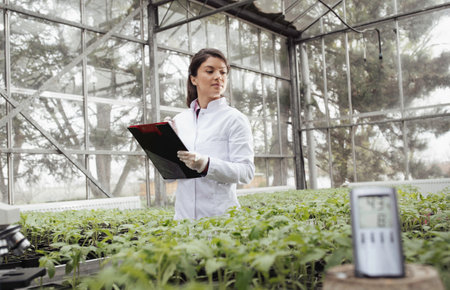 Pretty young woman agronomist writing notes about seedlings growth in greenhouse. Plant protection...
