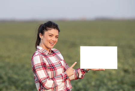 Pretty young farmer woman holding white empty board and pointing in it with finger, in field with...