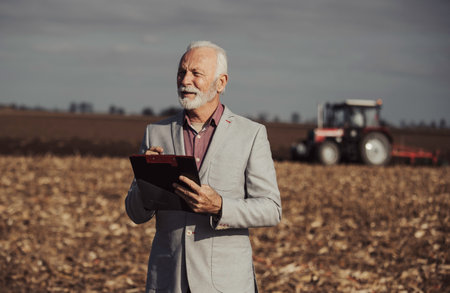 Portrait of mature businessman in suit standing on agricultural field in front of tractor in autumn timeの写真素材