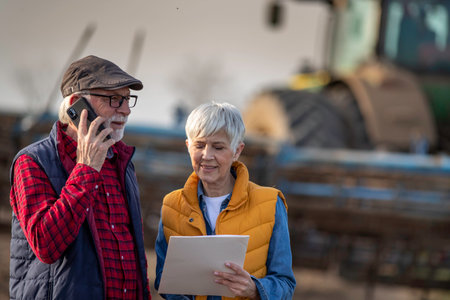 Mature farmers man and woman standing in field, writing notes and talking on mobile phone while...