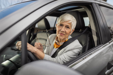 Senior woman with short silver hair smiles while holding her glasses in car. She is wearing stylish...