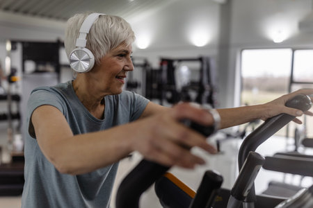 Senior woman enjoying workout on elliptical machine while listening to music through headphones in a...