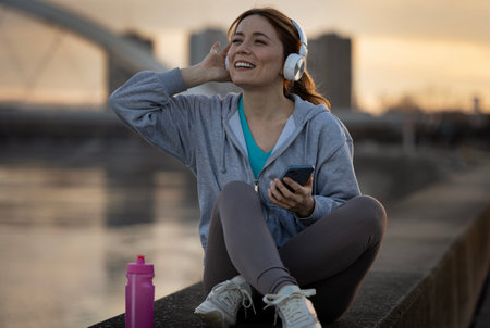 Cheerful woman sitting by riverside after workout, listening to music from mobile phone at sunset