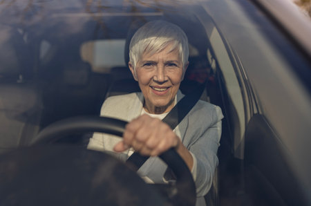 Cheerful elderly woman grips the steering wheel with enthusiasm while driving through a sunny landscape, showcasing her confidentの写真素材