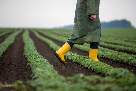 Agricultural worker in green raincoat and yellow gumboots walking through field in spring timeの写真素材