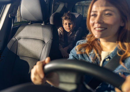 Woman smiling while driving car, with child seated comfortably in backseat. The sunlight brightens...