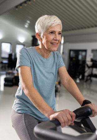 Female with gray hair enjoying cycling on stationary bike in well-lit gym, focusing on fitness and...