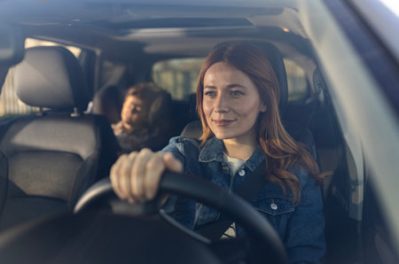 Woman smiling while driving car, with child seated comfortably in backseat. The sunlight brightens...