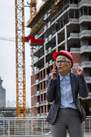 Business woman in gray suit and red hard hat speaking on phone at construction site, managing the ongoing project with focusの写真素材