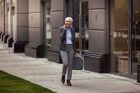 Business woman walking through busy city street, and having phone conversation. Modern buildings and people in the backgroundの写真素材