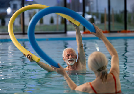 Senior man and woman holding pool noodles over their heads and exercising aqua aerobic in indoor...
