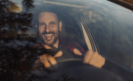 Man driving car, smiling while gripping the steering wheel. Bright trees and a sunny day create a...