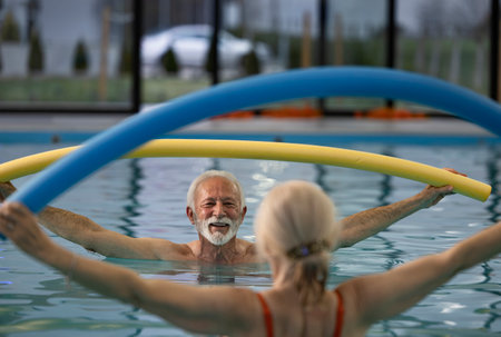 Senior man and woman holding pool noodles over their heads and exercising aqua aerobic in indoor...