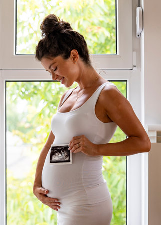 Pretty pregnant woman posing in front of a window as she holds both the ultrasound image and her big...