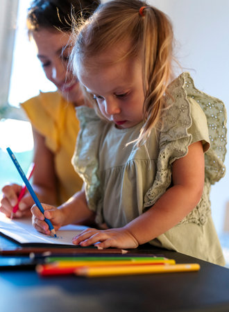 Mother and her young daughter engage in creative drawing session indoors during the evening,...