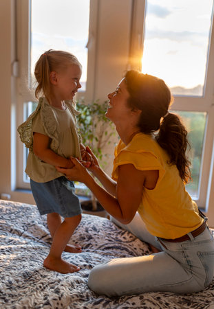 Side profile of mother and daughter joyfully laughing while playing on a bed in the evening, sunset...