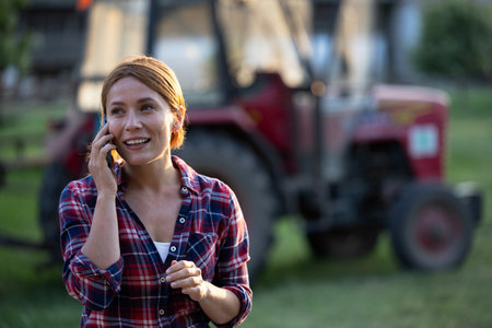 Young female farmer taking phone call with tractor in background on ranch, talking excitedly