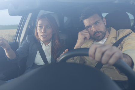 Frustrated man behind a steering wheel leaning his head on hand while woman gestures at road in...