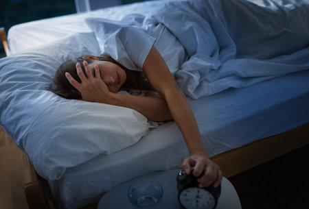 A woman lies in bed reaching for an alarm clock unhappy for waking early, while a glass of water sits on a table nearby. Morning light fills the room.の写真素材
