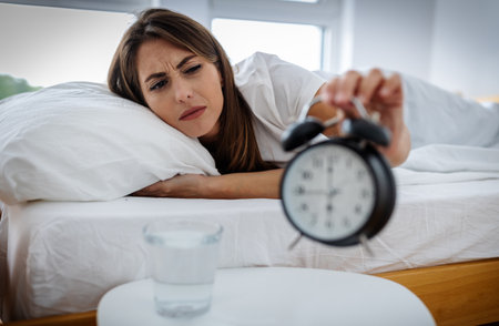 A woman lies in bed reaching for an alarm clock unhappy for waking early, while a glass of water sits on a table nearby. Morning light fills the room.の写真素材