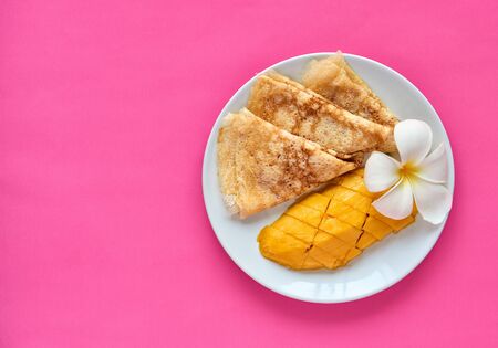 A slice of mango and homemade pancakes for breakfast. Decoration with a tropical flower. White plate on a pink background. Free space for text.の写真素材