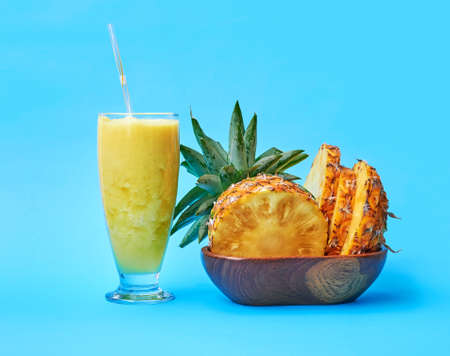 Jar filled with pineapple smoothies and a wooden plate with pineapple slices on a blue background
