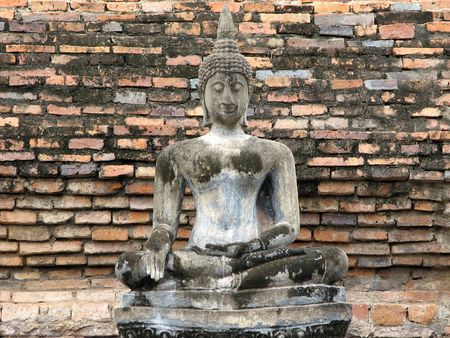A seated concrete Buddha statue in front of an ancient brick wall in sukhothai National Park.  Sukhothai, Thailand.の写真素材
