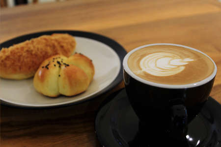 A glass of coffee latte with floral motifs on the coffee surface side by side with pastries on the tableの写真素材