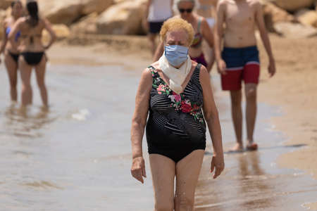 Torrenostra, CastellÃ³n, Spain - July 19, 2020: An elderly woman, retired, walks along the seashore wearing a mask to prevent the spread of COVID-19.のeditorial素材