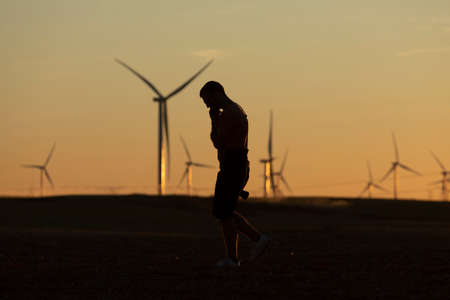 A man walks talking on the phone in the countryside at sunset, after taking pictures of distant wind turbines, on the horizon.の写真素材