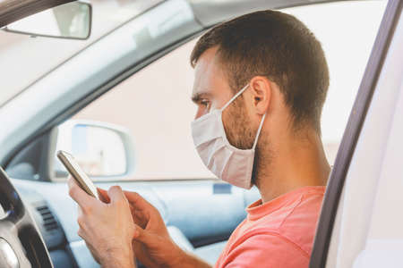 A young man, wearing a cloth face mask, checks the mobile phone inside the car, while he waits for...