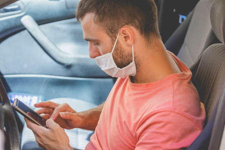 A young man, wearing a cloth face mask, checks the mobile phone inside the car, while he waits for...