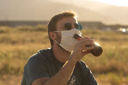A young man, with glasses, tries to drink a beer wearing a protective face mask.の写真素材