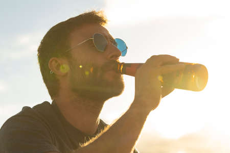 A young man, with glasses and a beard, drinks beer from a bottle, while enjoying the landscape and...