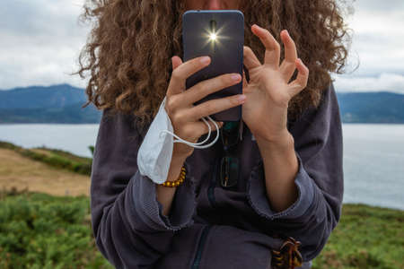 A young woman, grasping a protective face mask with her fingers, takes, at the same time, a photograph with a smart mobile phone, Spain.の写真素材