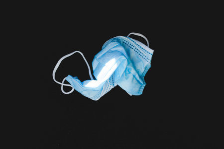 Protective face mask made of blue plastic, on a dark background, wrinkled, dirty and discarded,...