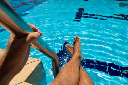 The feet of a young man, sitting on the edge of a pool filled with crystal clear blue water, cooling...