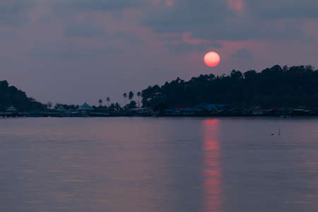 A huge sun, tinted red, sets behind the rainforest that surrounds the small town of Bang Bao, on the island of Koh Chang, Gulf of Thailandの写真素材