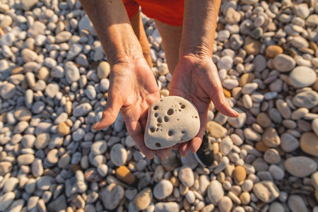 A woman, in her 70s, holds a stone full of holes, on a natural beach, next to the Mediterranean Sea,...
