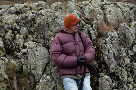 A mountaineer, senior adult man, in his 70s, in a feather coat, enjoys the scenery while resting leaning on some rocks full of lichen, Pyrenees, Spainの写真素材