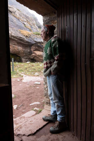 A mountaineer, senior adult man, in his 70s, with handmade woolen sweater, observes the outside from the door of a mountain hut, Pyrenees, Spainの写真素材