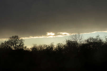 A winter landscape of clouds and dark silhouettes of bare trees, at sunset, from a hotel window near Manchester Airportの写真素材